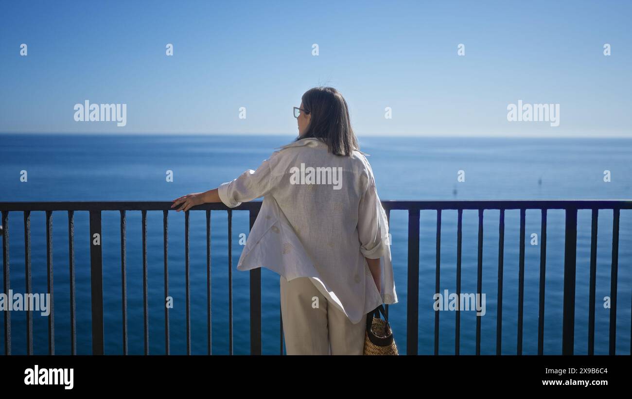 Giovane donna ispanica che gode della vista panoramica dell'oceano da un punto di osservazione a polignano a mare, puglia, italia, sotto cieli azzurri. Foto Stock