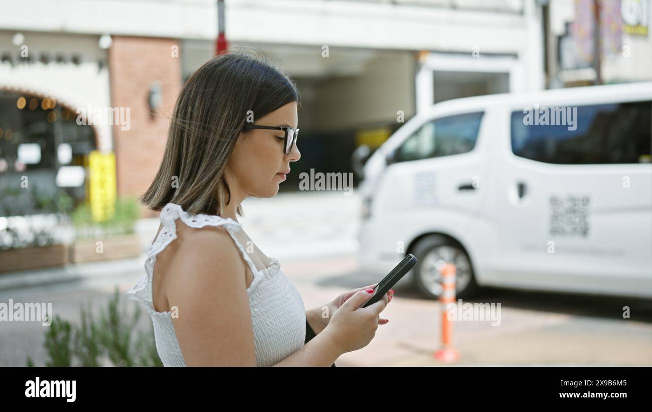 Bella donna ispanica con gli occhiali che cammina con sicurezza per le strade di tokyo mentre è coinvolta nel suo telefono, scrivendo un serio messaggio di lavoro Foto Stock