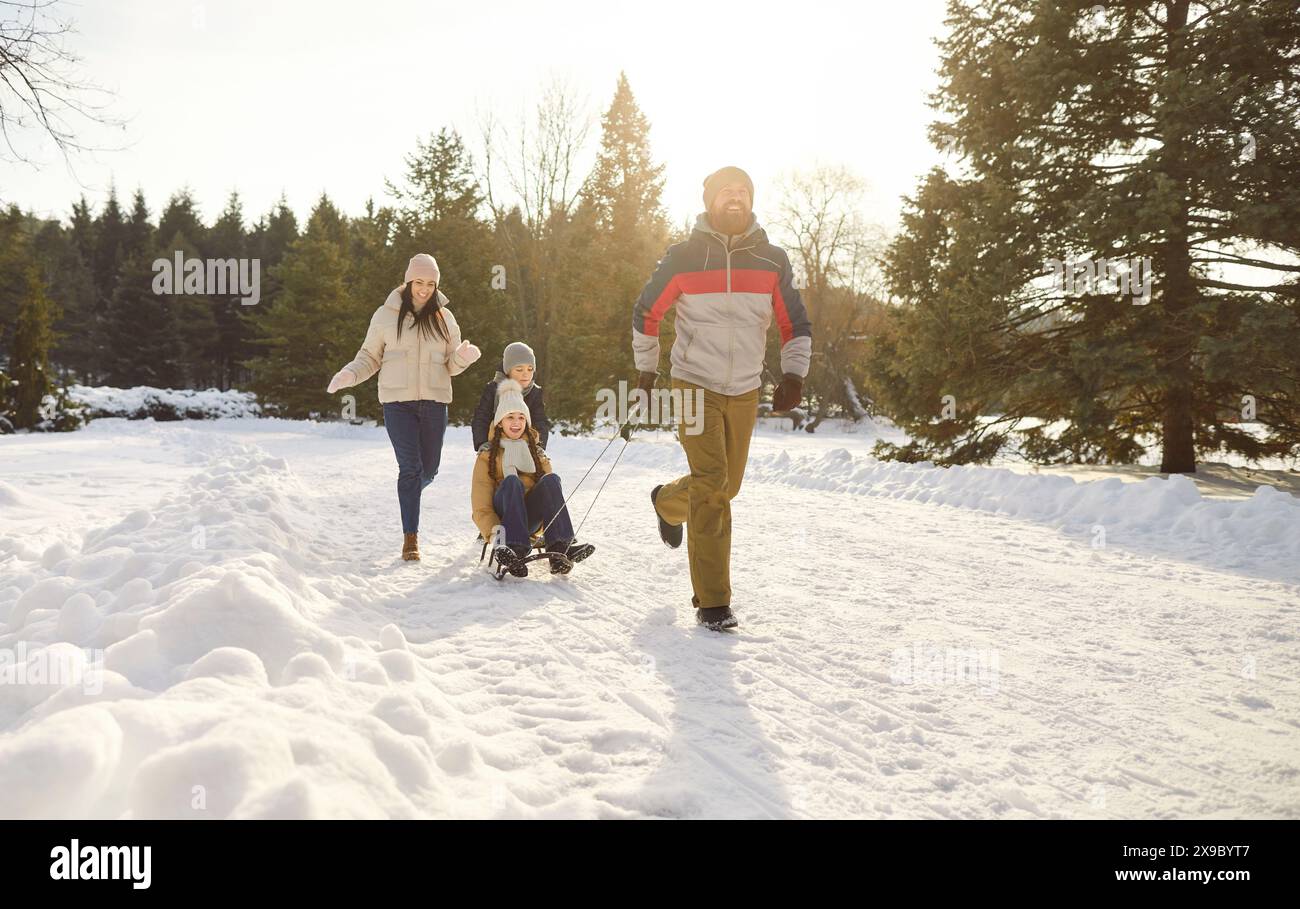 I genitori portano i bambini sulle slitte mentre camminano felicemente lungo i sentieri innevati nel parco invernale. Foto Stock