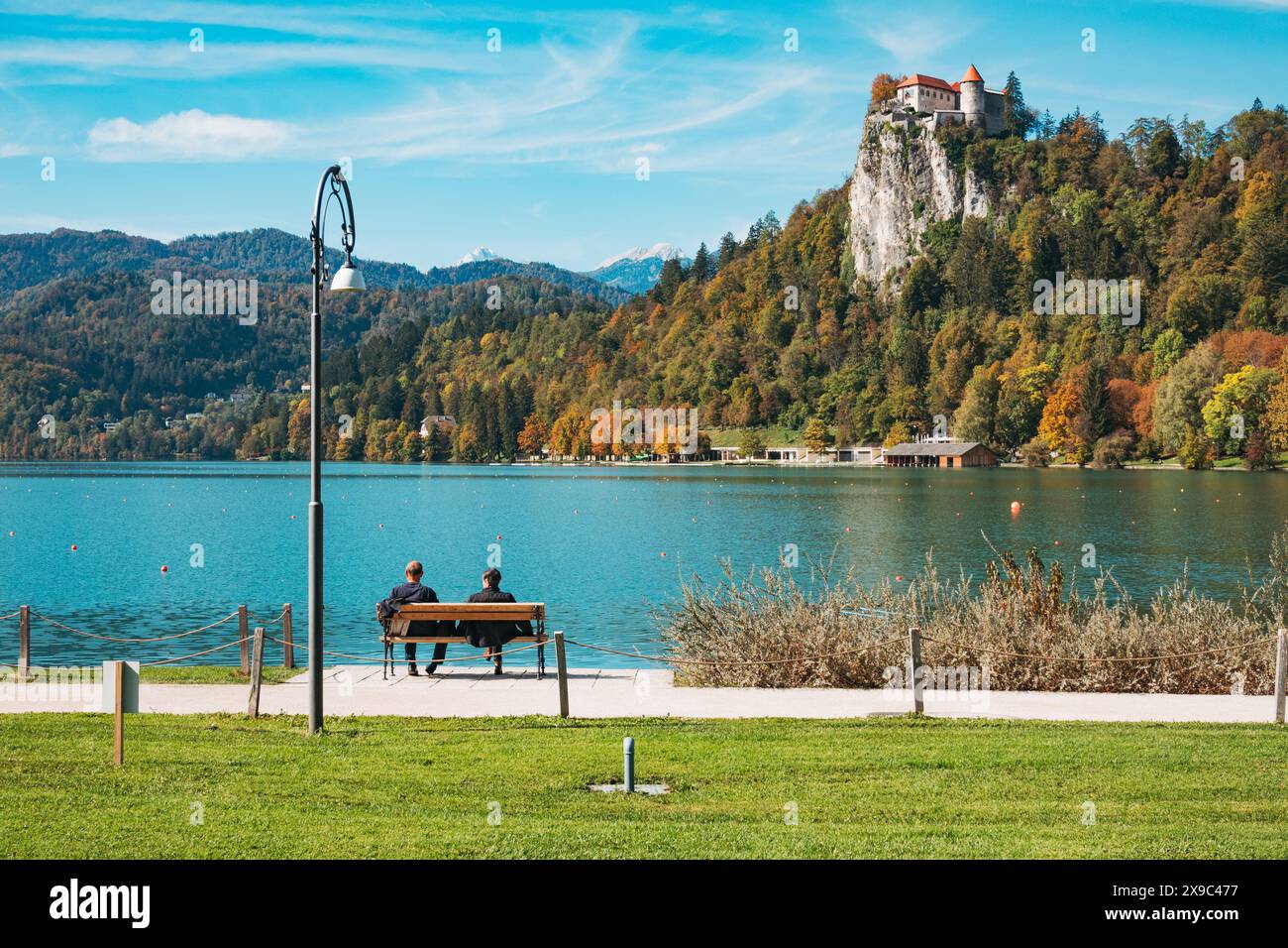 Due persone siedono su una panchina che guarda il castello di Bled, arroccato sulla cima di una scogliera vicino al lago Bled, in una giornata di autunno limpida Foto Stock