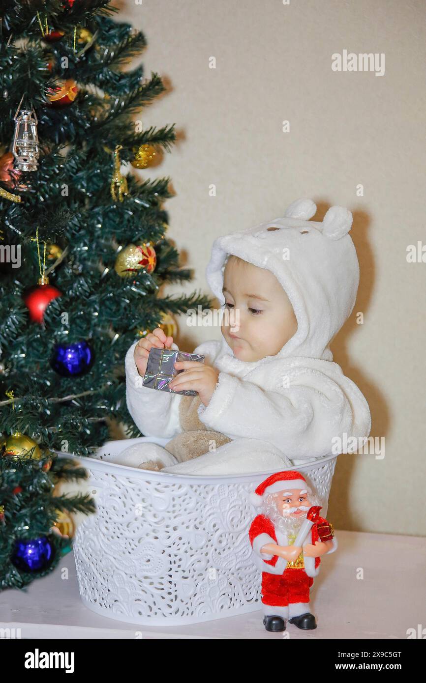 Un bambino in abiti bianchi si siede in un cestino bianco accanto a un albero di Natale decorato e a una statuetta di Babbo Natale Foto Stock