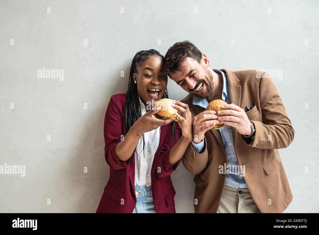 Due giovani hipster espatriano uomini d'affari che si uniscono a mangiare cibo poco salutare o cibo di strada, tenendo in mano due hamburger con grasso e grasso Foto Stock