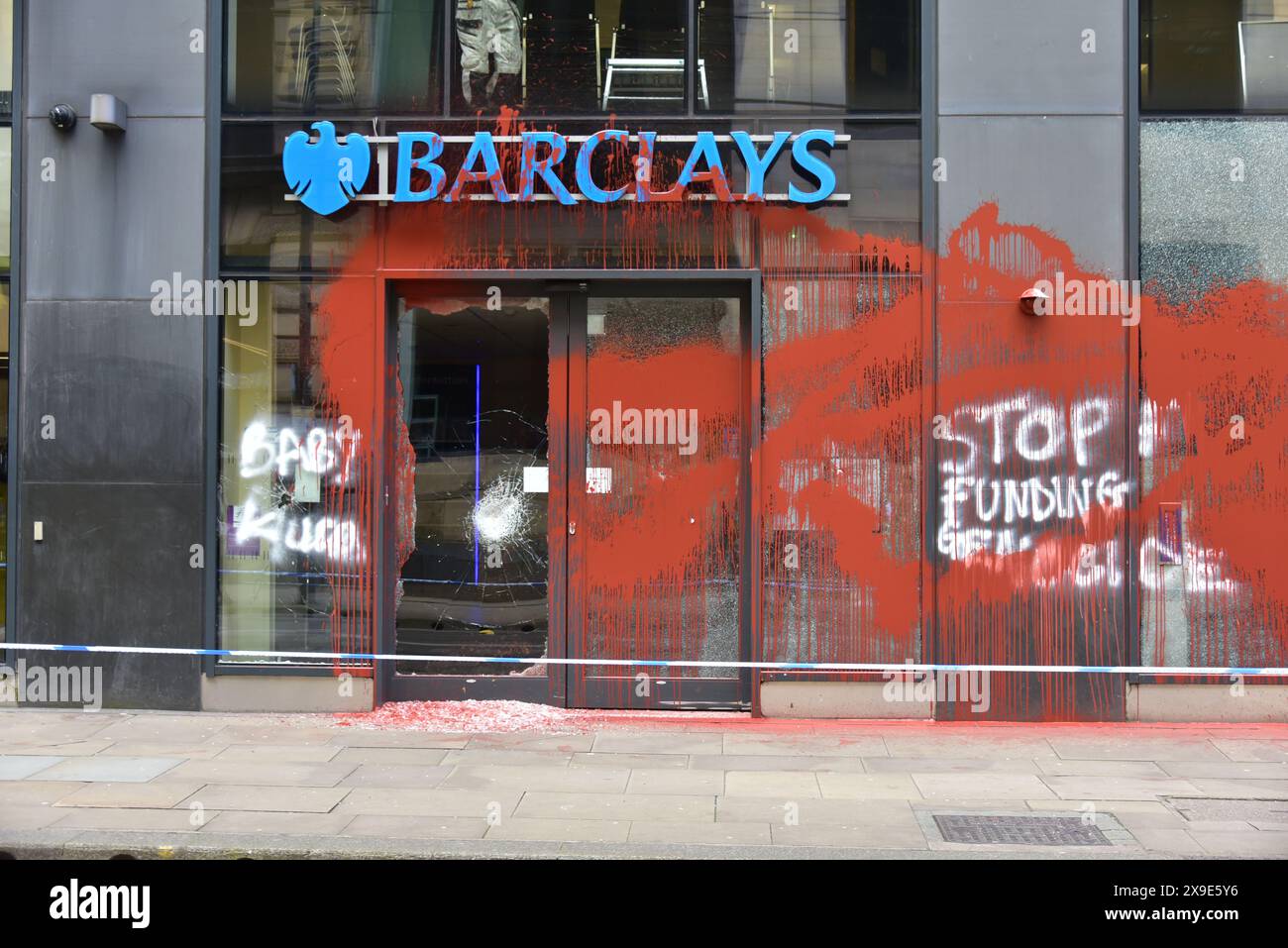 Barclays Bank, Mosley Street, centro di Manchester, Regno Unito, una porta di vetro si è rotta e coperta di vernice rossa il 31 maggio 2024. La polizia ha messo in atto un cordone. Negli ultimi mesi i manifestanti pro-Palestina hanno preso di mira questa banca e gettato vernice rossa ad altre imprese che credono stiano sostenendo Israele. I graffiti sul fronte includono: "Smettete di finanziare il genocidio”. Foto Stock