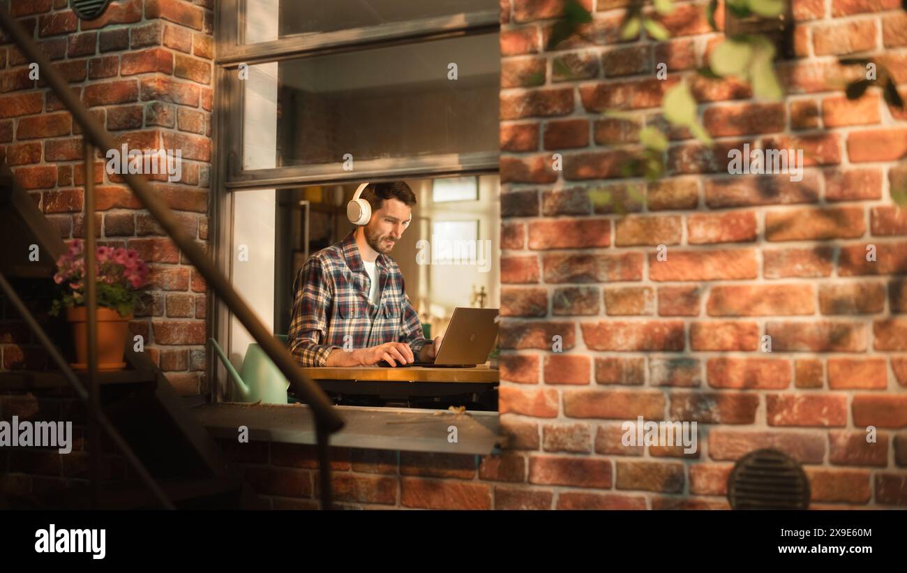 Moderna digitazione freelance su un notebook, ascolto di musica con le cuffie. Concetto di imprenditore e-Business professionale e di successo presso l'ufficio domestico. Vista dall'esterno verso la finestra. Foto Stock