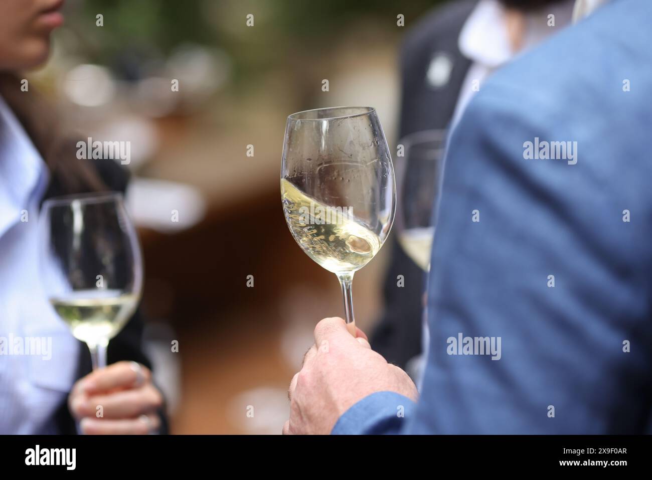 Profondità di campo bassa (messa a fuoco selettiva) dettagli con la mano di un uomo che tiene un bicchiere di vino bianco a una festa. Foto Stock