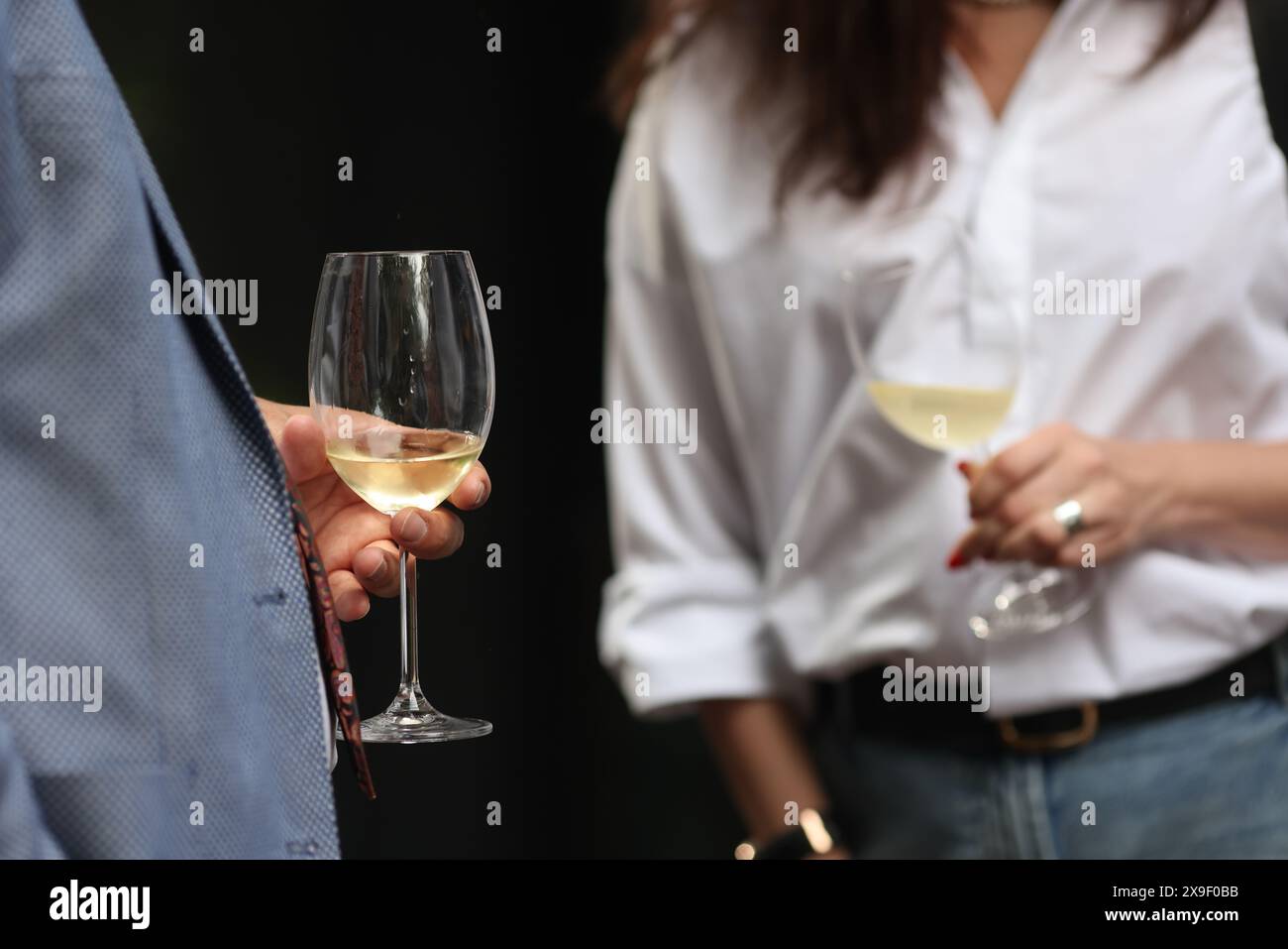 Profondità di campo bassa (messa a fuoco selettiva) dettagli con la mano di un uomo che tiene un bicchiere di vino bianco a una festa. Foto Stock