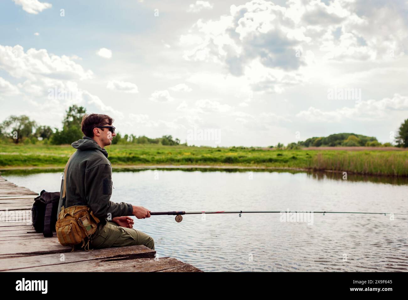 Ritratto a tutto il corpo di un giovane pescatore che indossa l'uniforme militare. Un tizio seduto sul ponte sul fiume tenendo la canna accanto allo zaino. Ricreazione e lui mentale Foto Stock