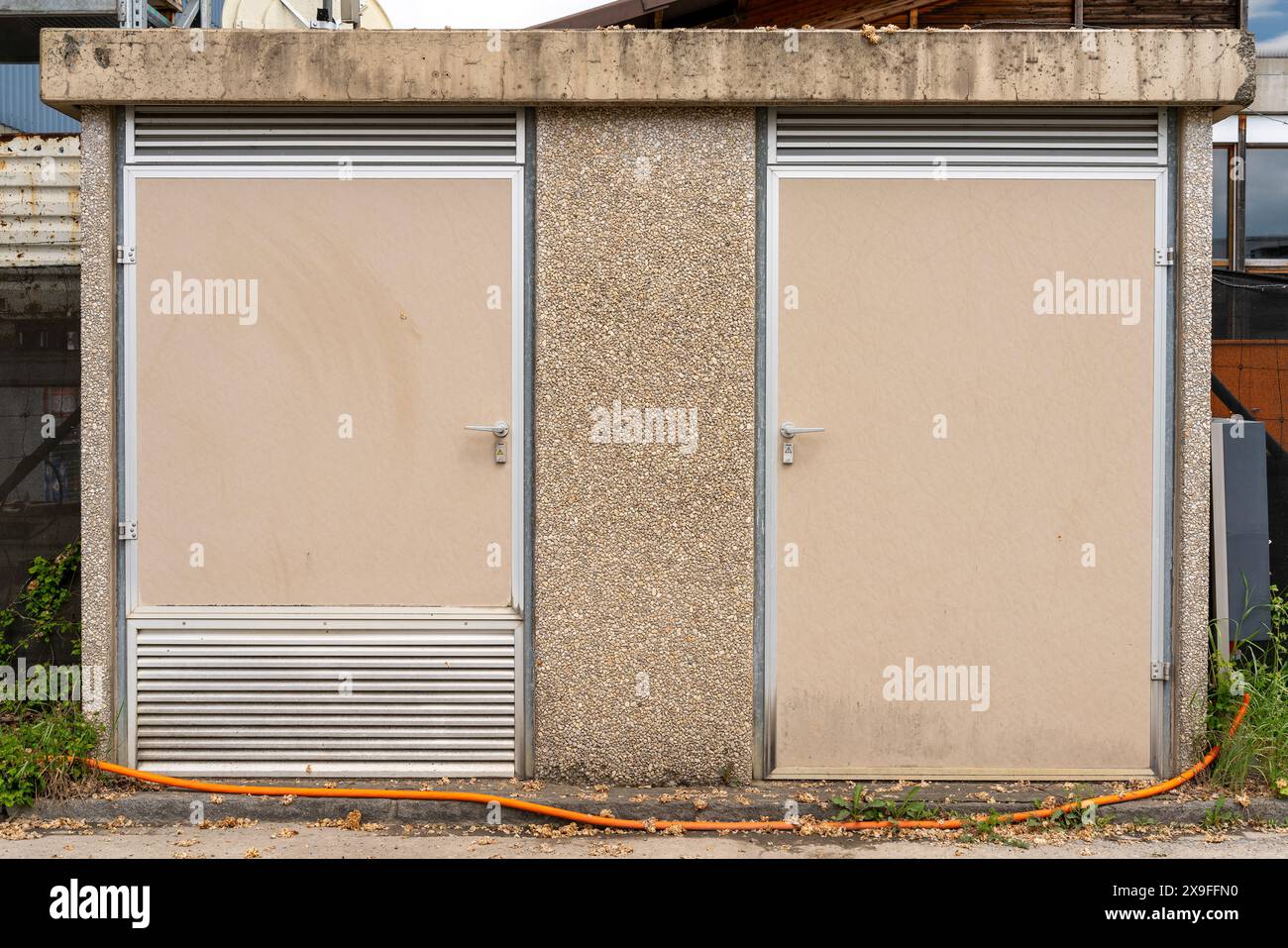 Casa trasformatore con due porte chiuse e piccoli segnali di avvertimento elettrici. Grande cavo arancione che corre davanti a quella stazione. Foto Stock