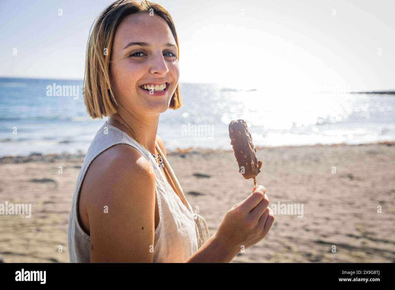 Una donna sorride e tiene in mano una bacchetta di gelato al cioccolato su una spiaggia Foto Stock