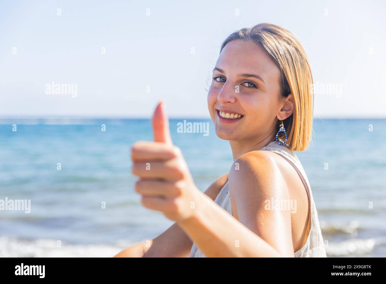 Una giovane donna rossa sorride e regala un pollice mentre è in piedi su una spiaggia Foto Stock