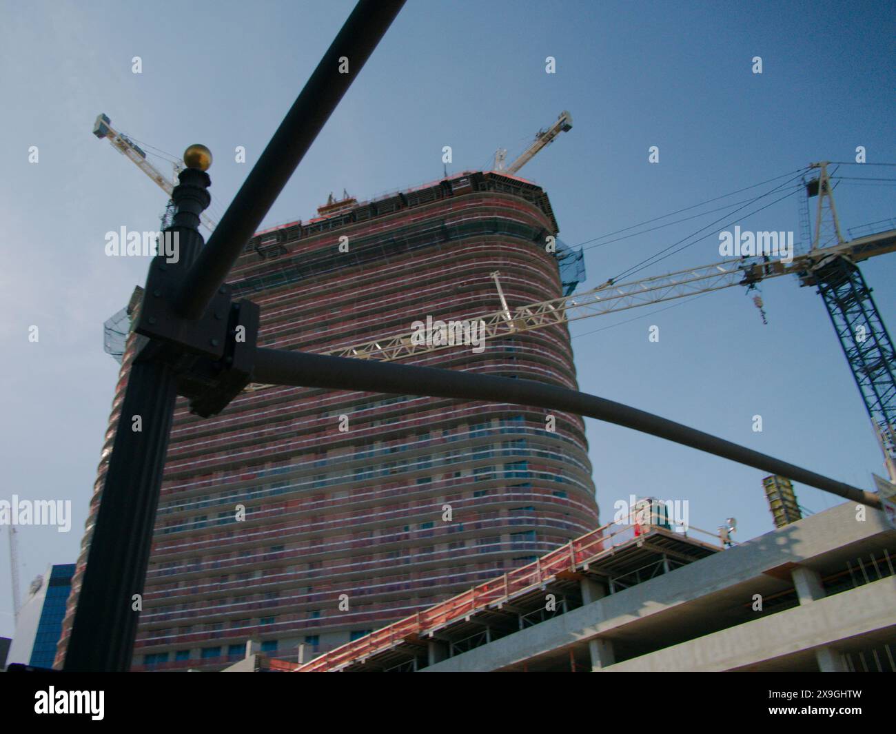 Ammira il palo in primo piano del parcheggio su più piani, l'edificio e le gru in costruzione. Cielo blu e recinzione arancione. Niente persone Foto Stock