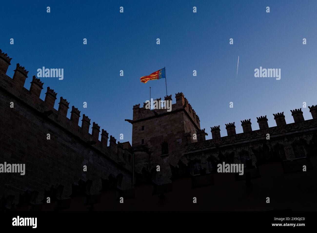 La bandiera della comunità valenciana ondeggia in cima a un edificio storico con merlature contro un cielo azzurro cristallino durante il festival las fallas Foto Stock