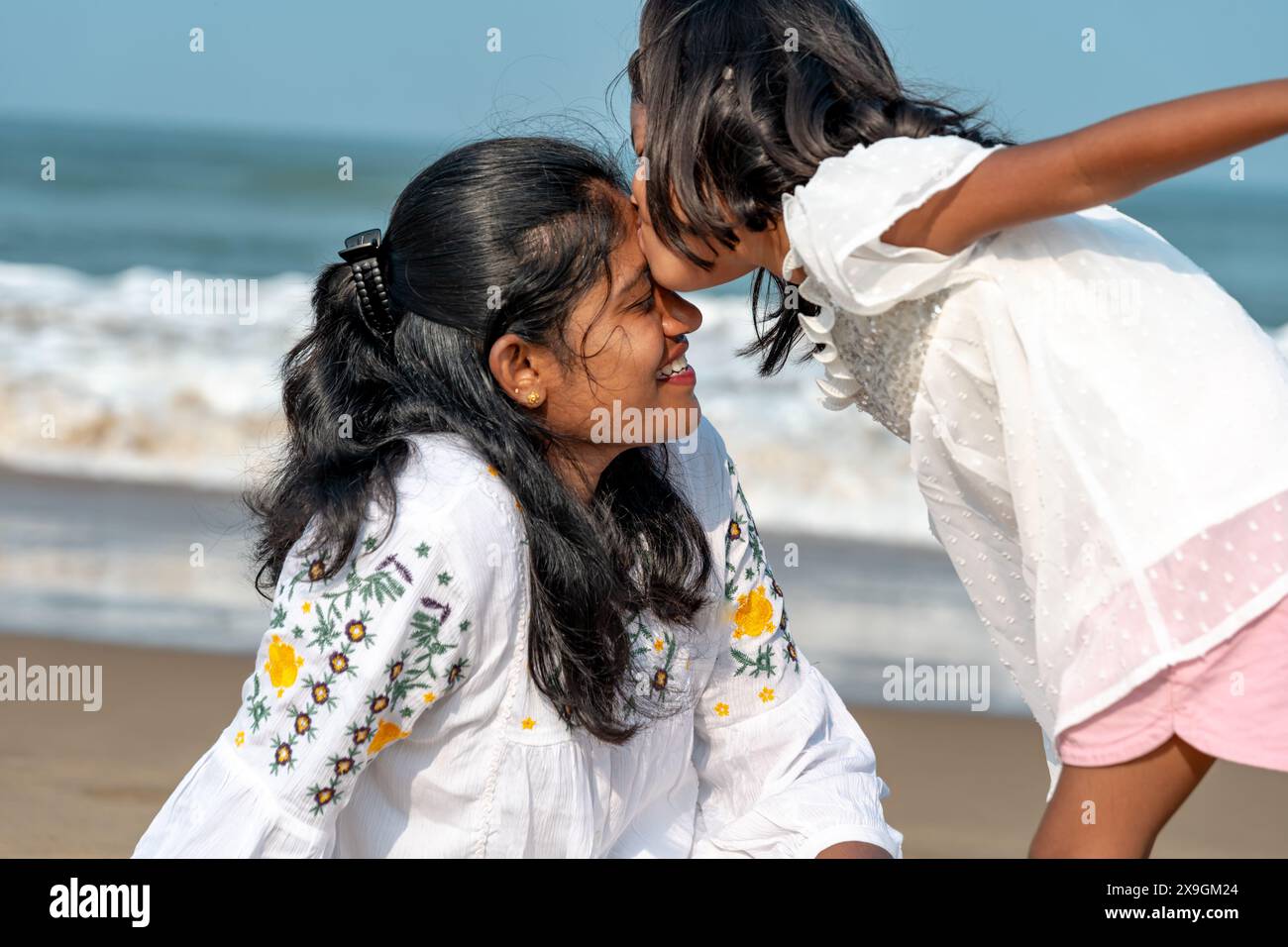 una giovane figlia con un vestito bianco si appoggia per baciare la fronte di sua madre su una spiaggia baciata dal sole. Il loro legame irradia amore e calore. Foto Stock