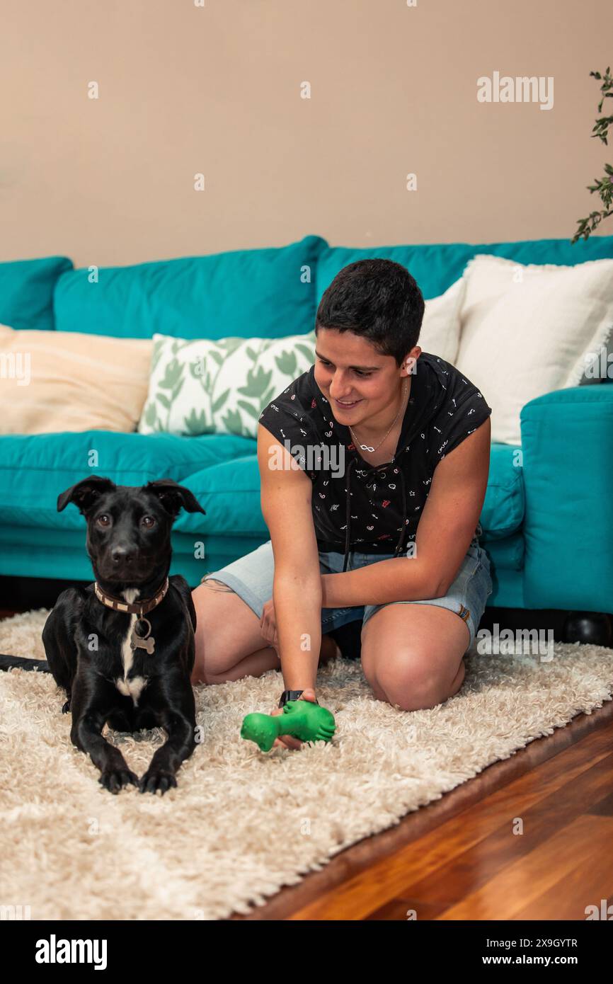 Vertical Una giovane donna giocosa con i capelli corti trascorre del tempo di qualità con il suo amato cane, partecipando a una divertente sessione di gioco sul pavimento accogliente della sua città Foto Stock