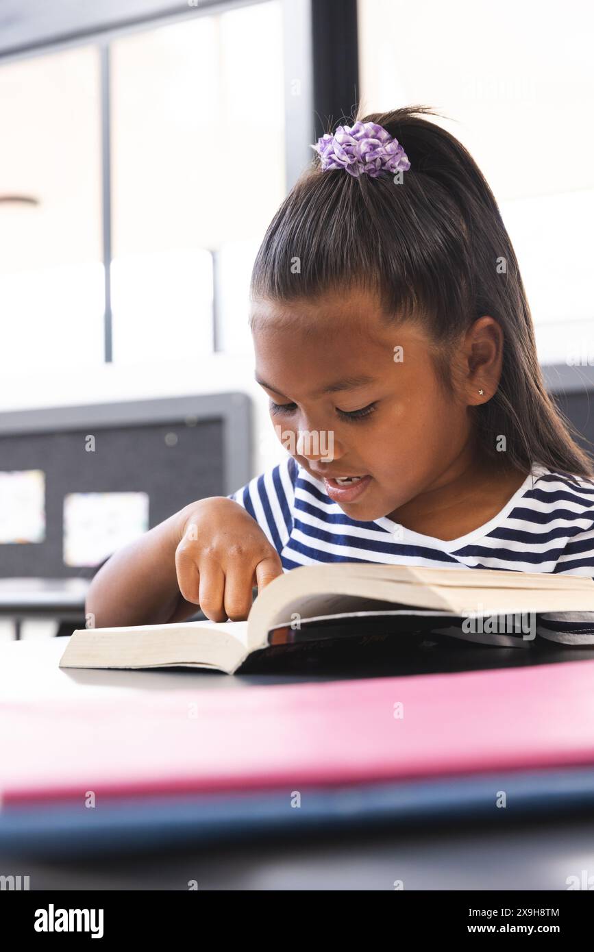 A scuola, giovane ragazza birazziale che si concentra sulla lettura di un libro in classe Foto Stock