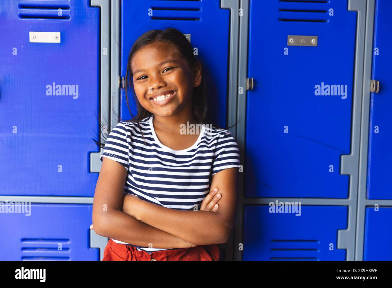 A scuola, la giovane studentessa birazziale si trova all'aperto, con le braccia incrociate, sorridendo Foto Stock