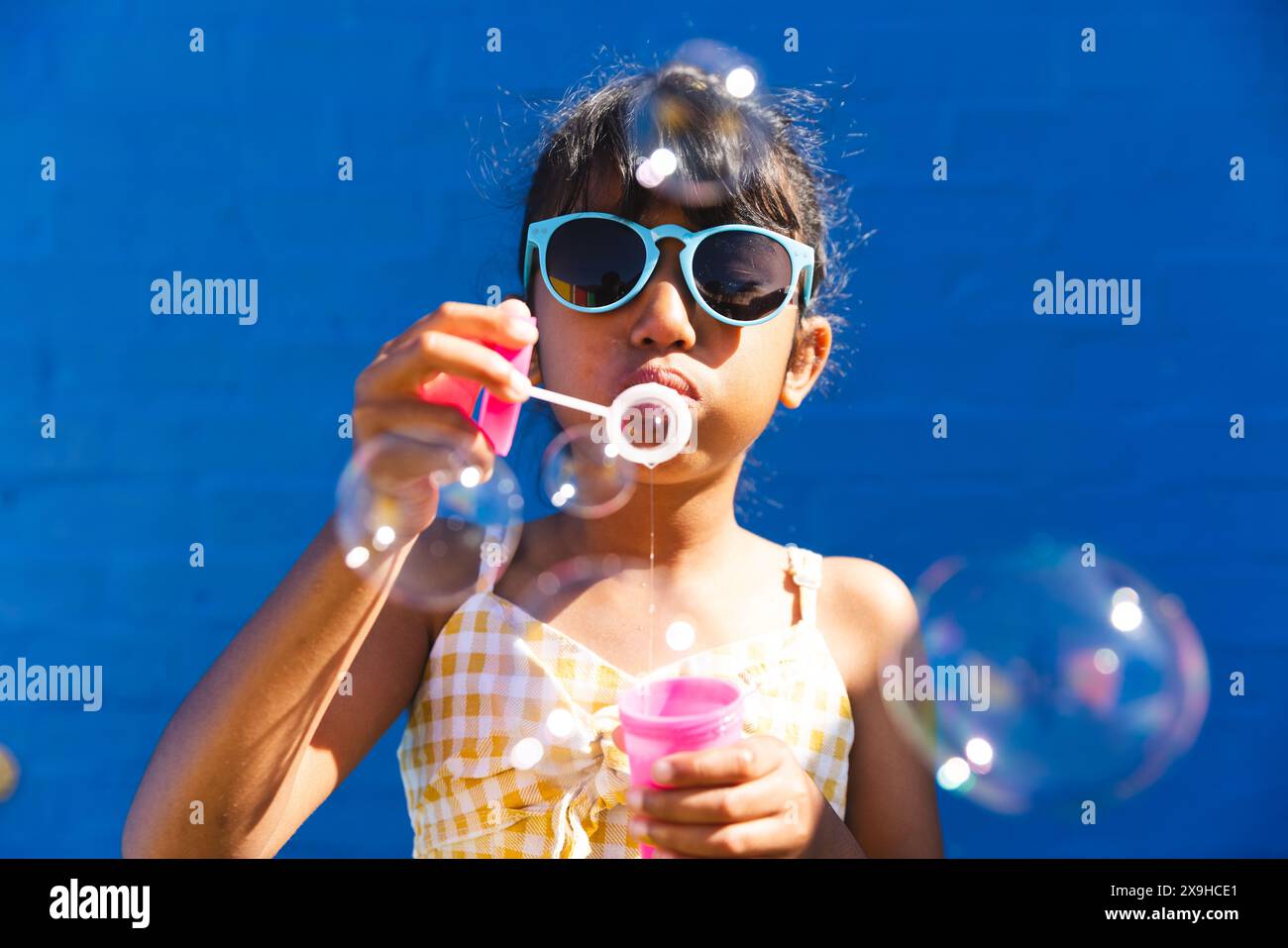 Una ragazza birazziale che indossa occhiali da sole e un abito da sole ama soffiare bolle all'aperto Foto Stock