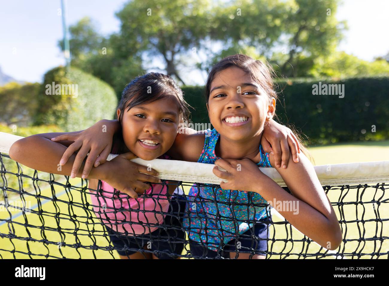 All'aperto, due giovani sorelle birazziali sorridenti e appoggiate su una rete da tennis nelle giornate di sole Foto Stock