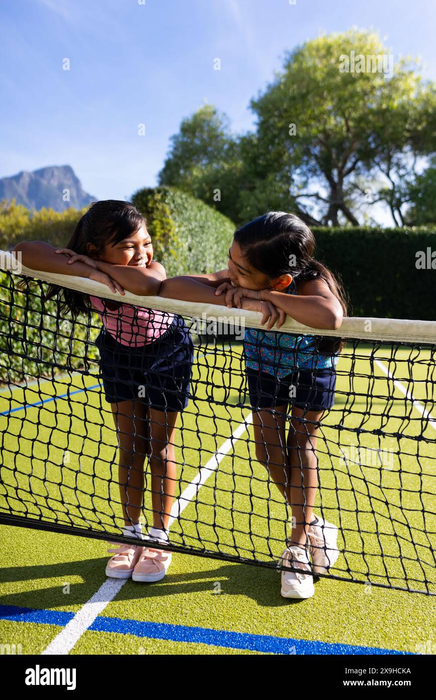 All'aperto, due giovani sorelle birazziali si appoggiano su una rete da tennis, sorridendo l'una all'altra Foto Stock