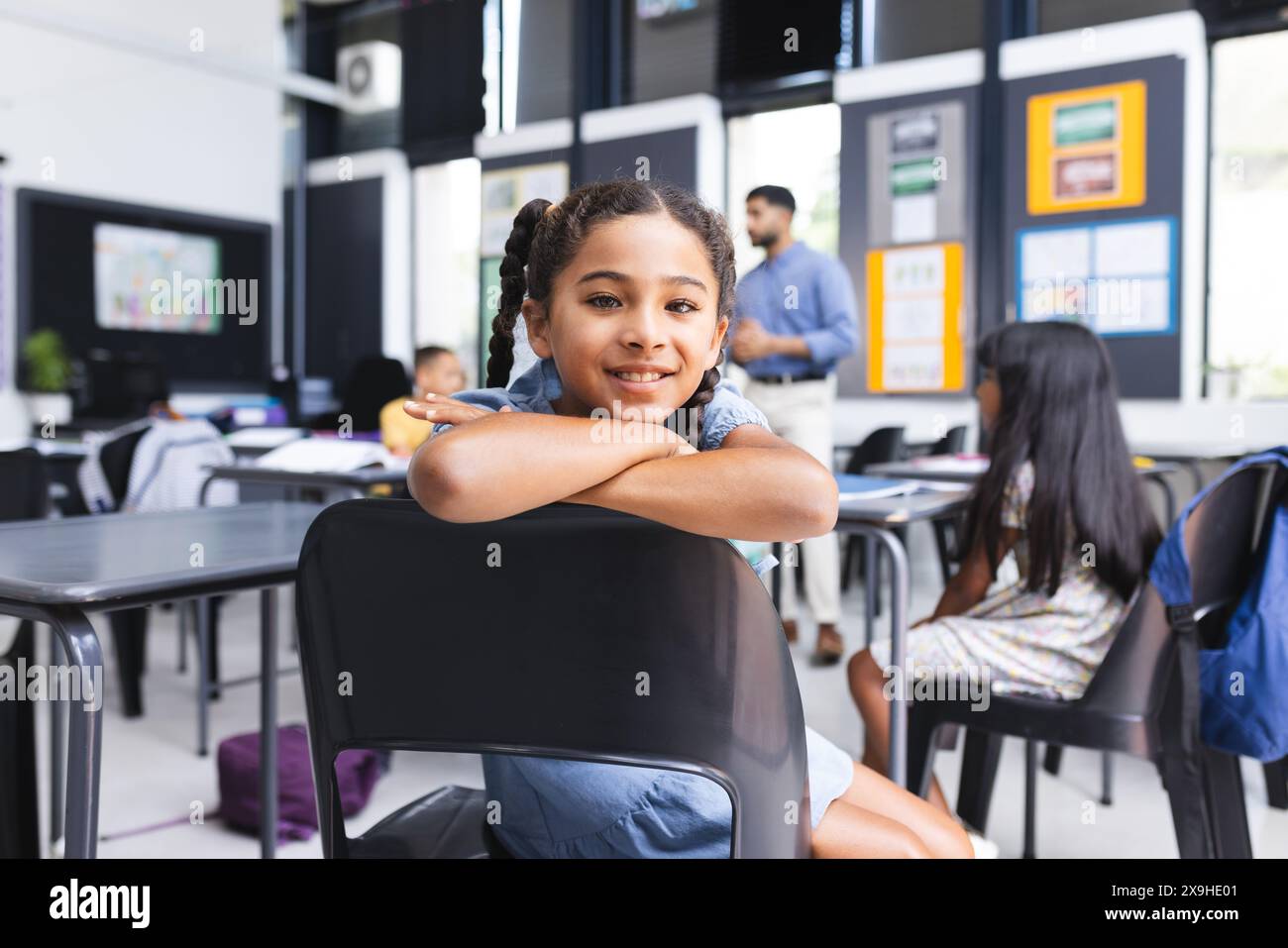 Ragazza birazziale con i capelli intrecciati sorride in un'aula scolastica. Insegnanti e studenti in background suggeriscono un ambiente educativo vivace. Foto Stock