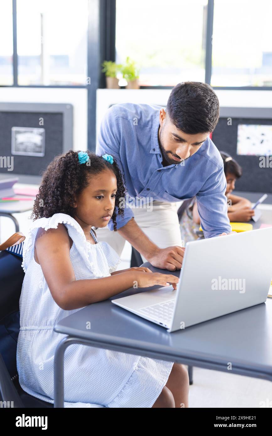 A scuola, insegnante asiatico maschile che aiuta la giovane studentessa birazziale con un portatile in classe Foto Stock
