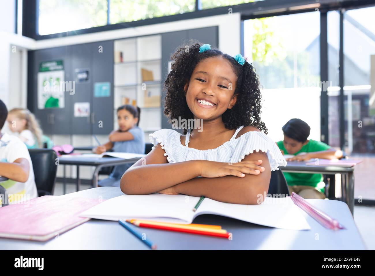 A scuola, la giovane ragazza birazziale con i capelli neri ricci è seduta ad una scrivania in classe, sorridente Foto Stock