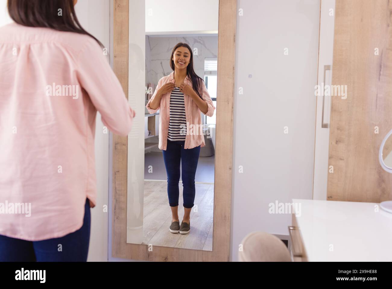 Una giovane donna birazziale a casa, che indossa abiti casual, sorride al suo riflesso Foto Stock