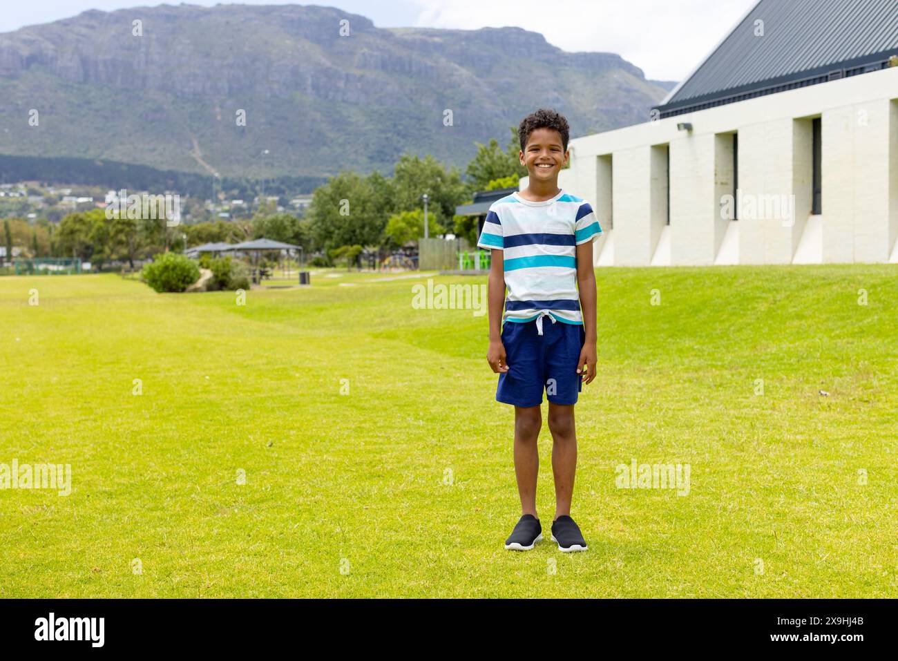 Il ragazzo birazziale sorride su un campo di erba soleggiata, montagne sullo sfondo, con spazio per copiare Foto Stock
