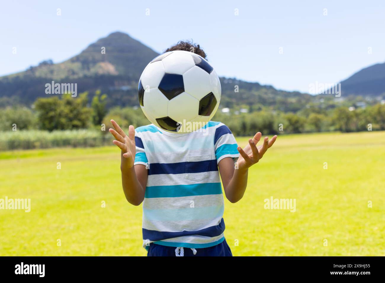 Un ragazzo birazziale gioca con un pallone da calcio all'aperto, circondato dal verde e dalle montagne Foto Stock