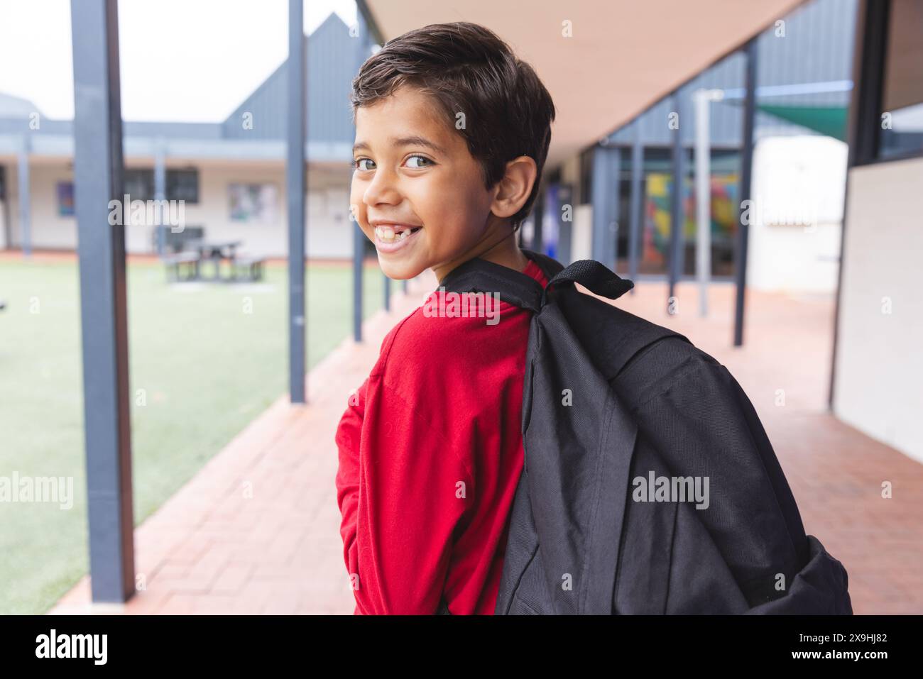 A scuola, il giovane studente birazziale maschile che indossa uno zaino sorride all'aperto Foto Stock