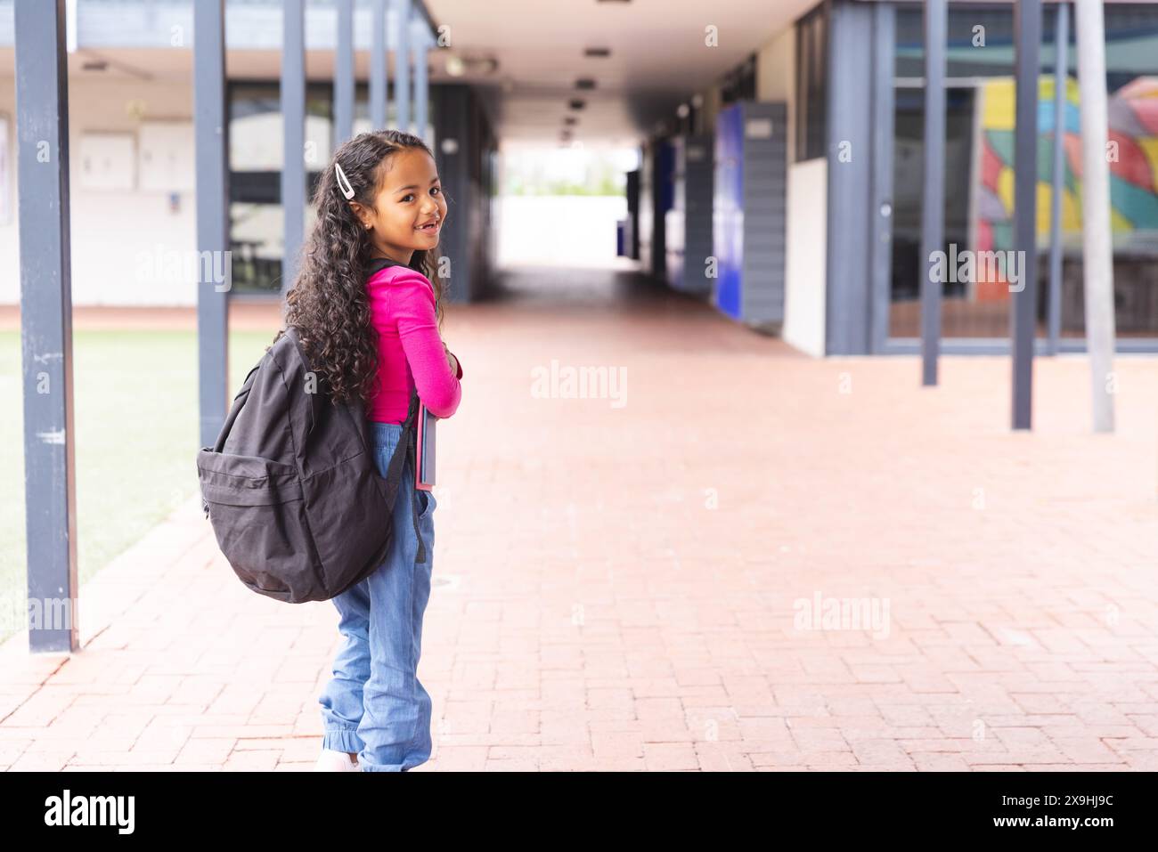A scuola, una giovane ragazza birazziale che indossa uno zaino in piedi in un corridoio all'aperto con spazio per le copie Foto Stock