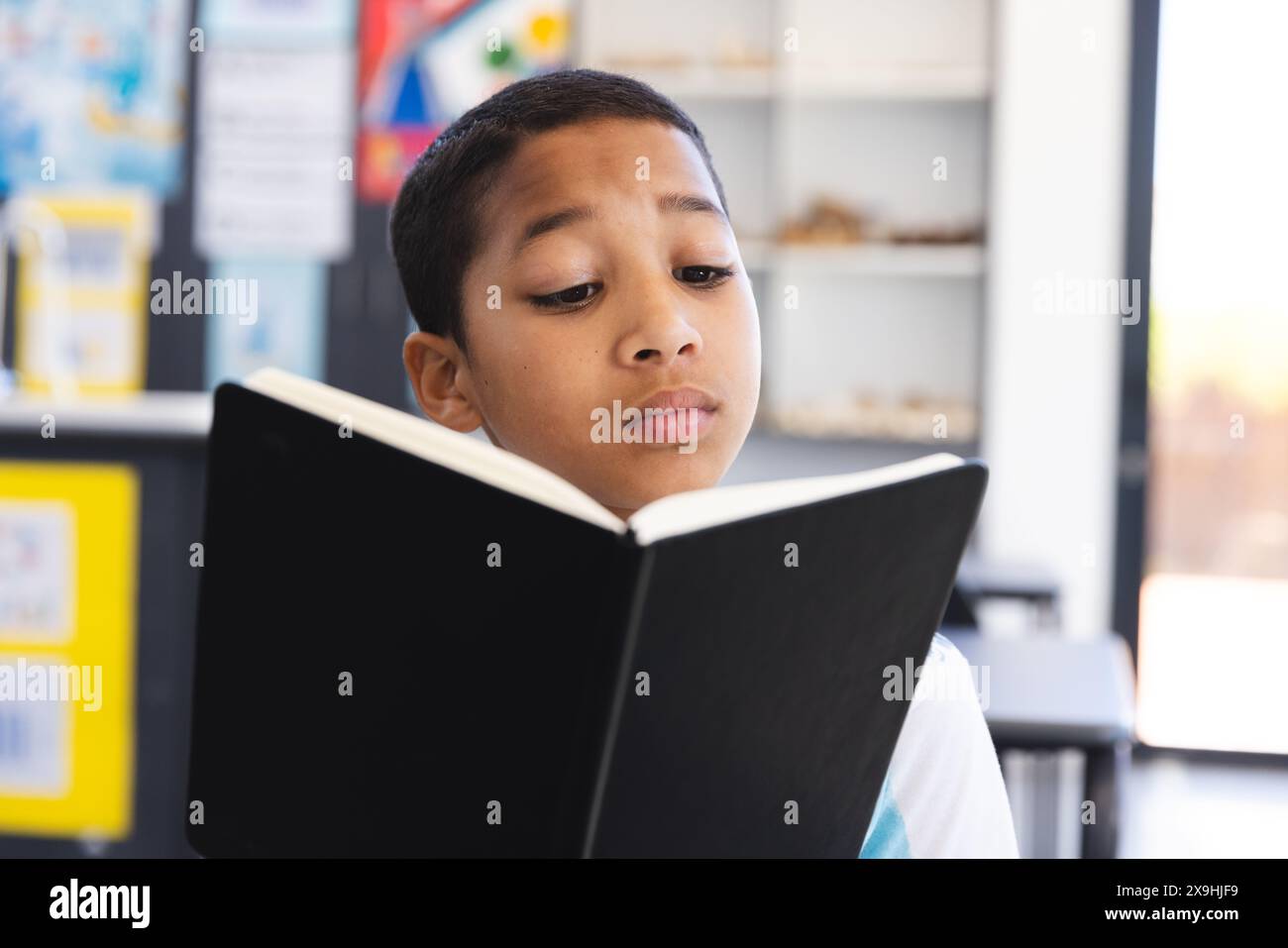 Ragazzo birazziale impegnato a leggere un libro in classe a scuola. Si concentra sull'apprendimento, circondato da un ambiente luminoso in classe. Foto Stock