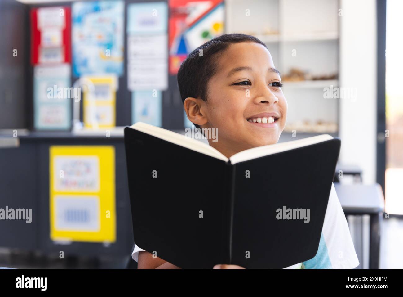 Un ragazzo birazziale ama leggere un libro in classe a scuola. Il suo sorriso luminoso riflette la gioia di imparare in un ambiente educativo. Foto Stock