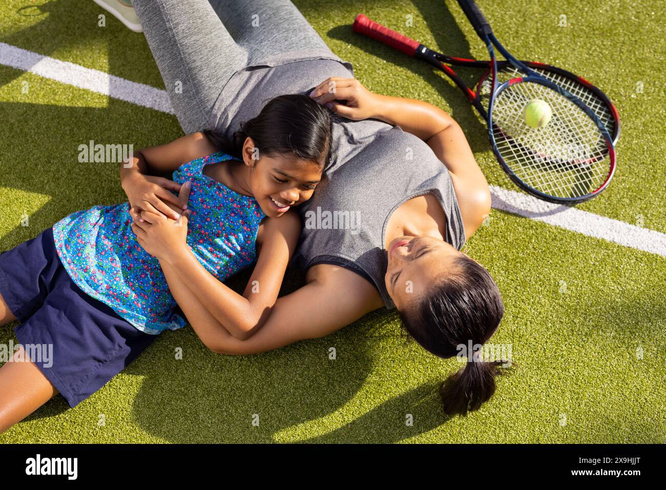 All'aperto, madre e bambino birazziali giacciono sull'erba, sorridono e abbracciano dopo il tennis Foto Stock