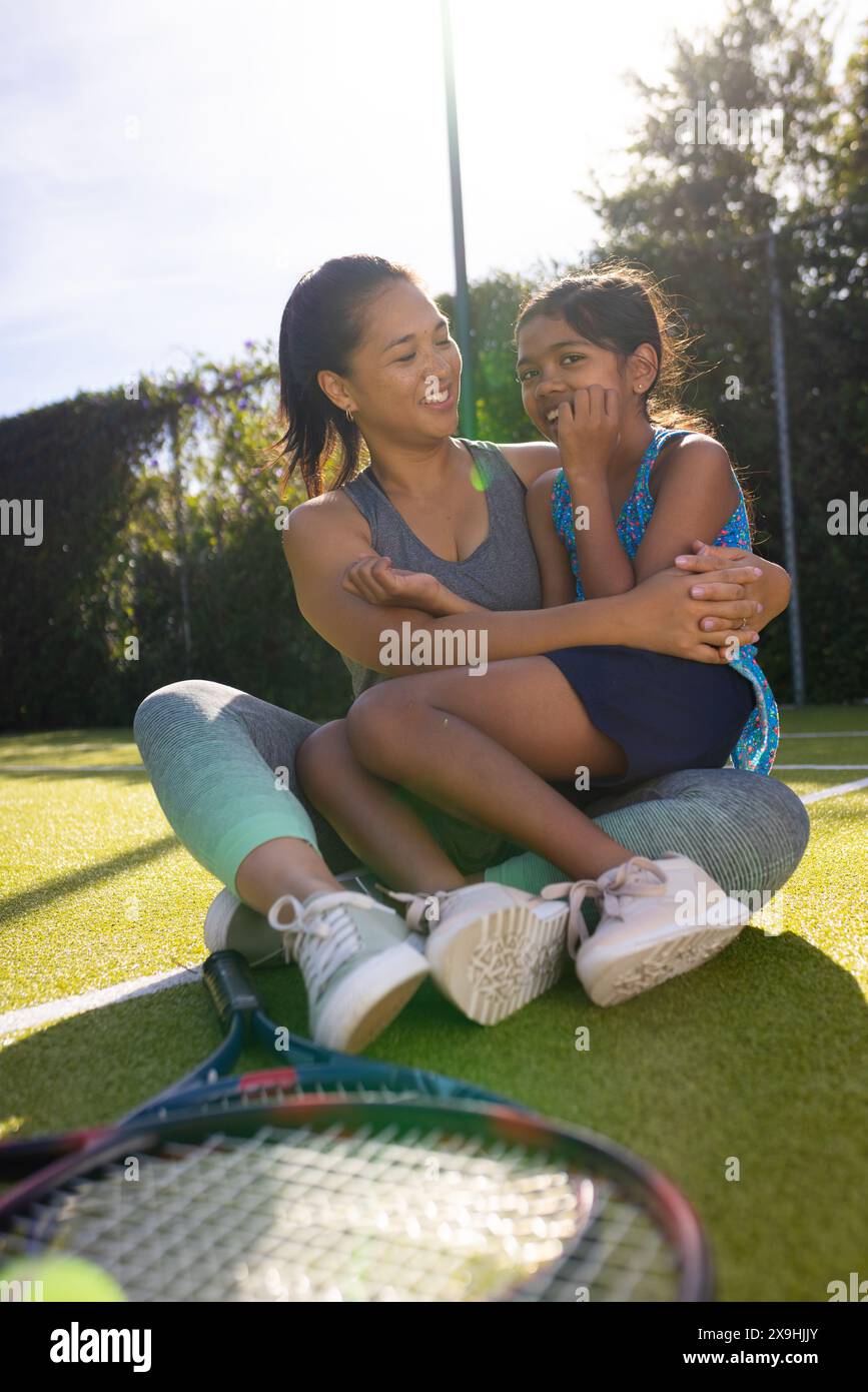 All'aperto, madre e figlia birazziali siedono sul campo da tennis, sorridendo e abbracciando Foto Stock