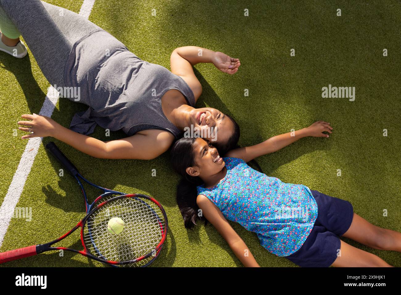 All'aperto madre e figlia birazziali sdraiate sull'erba con racchette da tennis, sorridenti Foto Stock