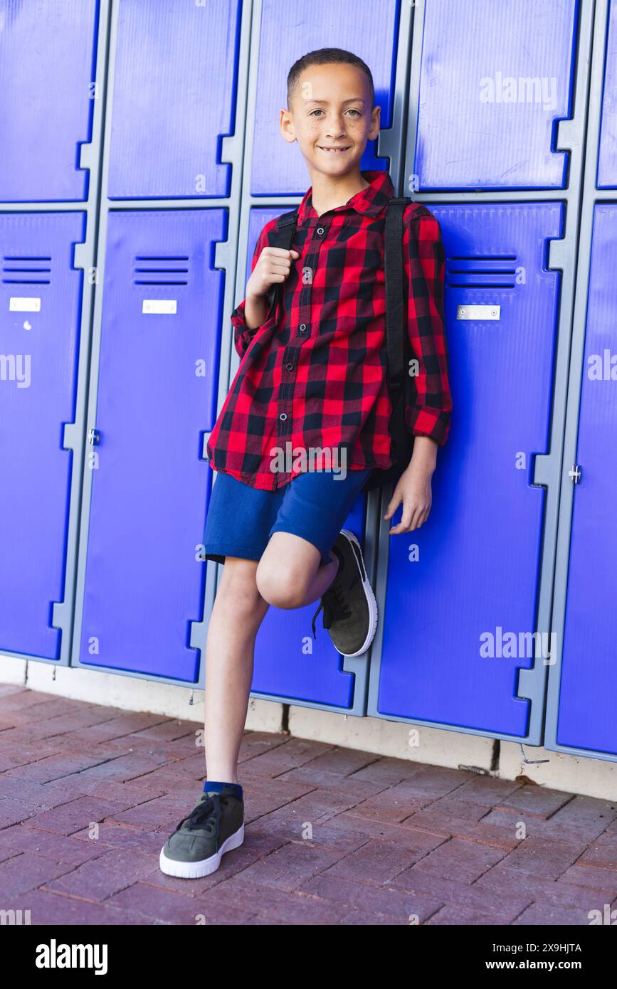 Il ragazzo birazziale si appoggia contro gli armadietti blu a scuola, indossando una camicia a scacchi rossa e nera Foto Stock