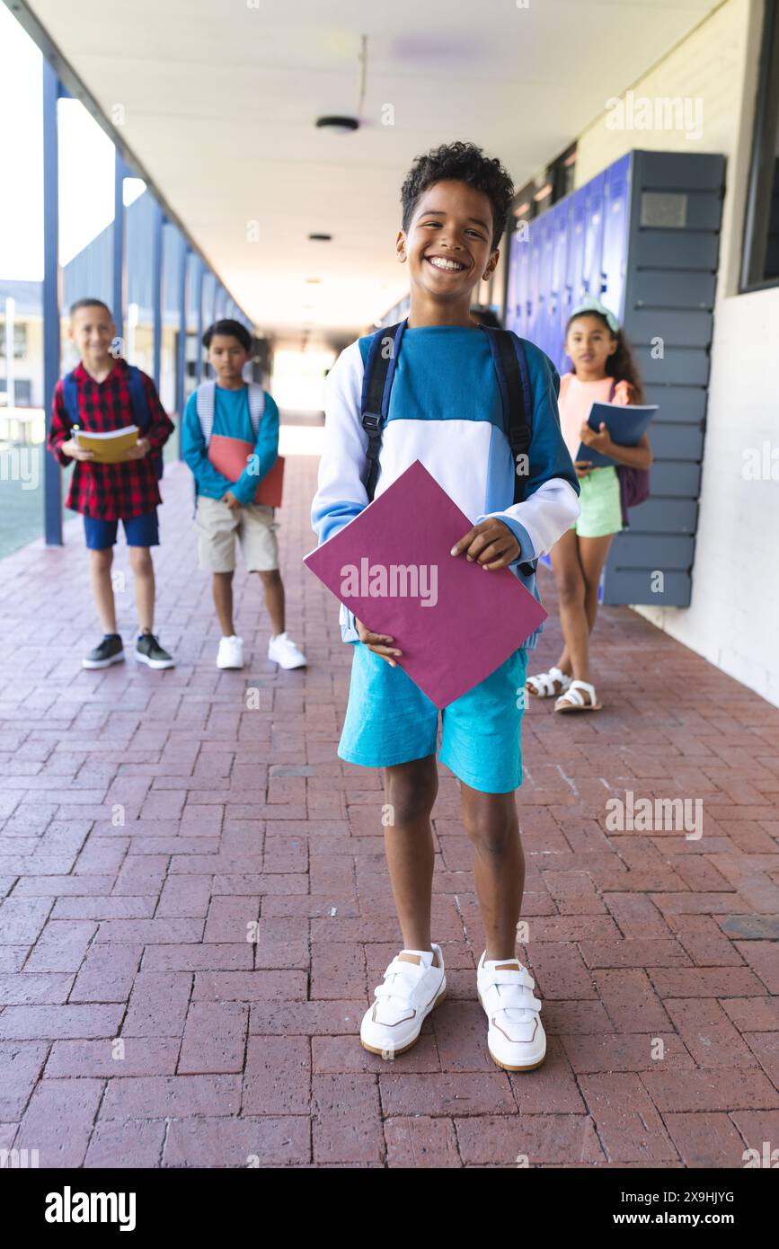 Un ragazzo birazziale in primo piano sorride, tiene una cartella, con compagni di classe a scuola Foto Stock