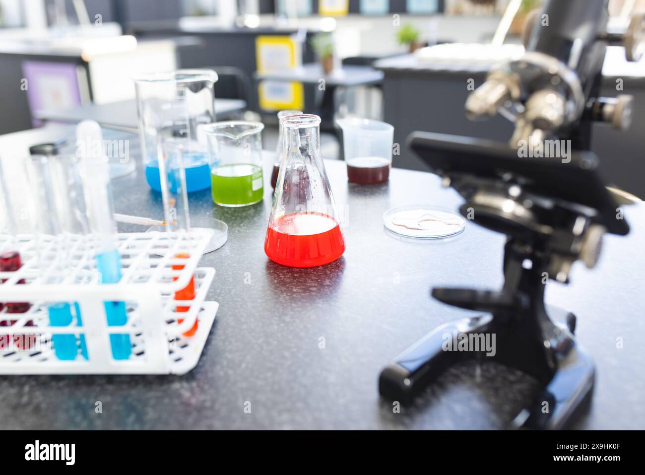 Un microscopio si trova accanto a becher colorati riempiti di liquido e provette per analisi su un banco da laboratorio a scuola Foto Stock