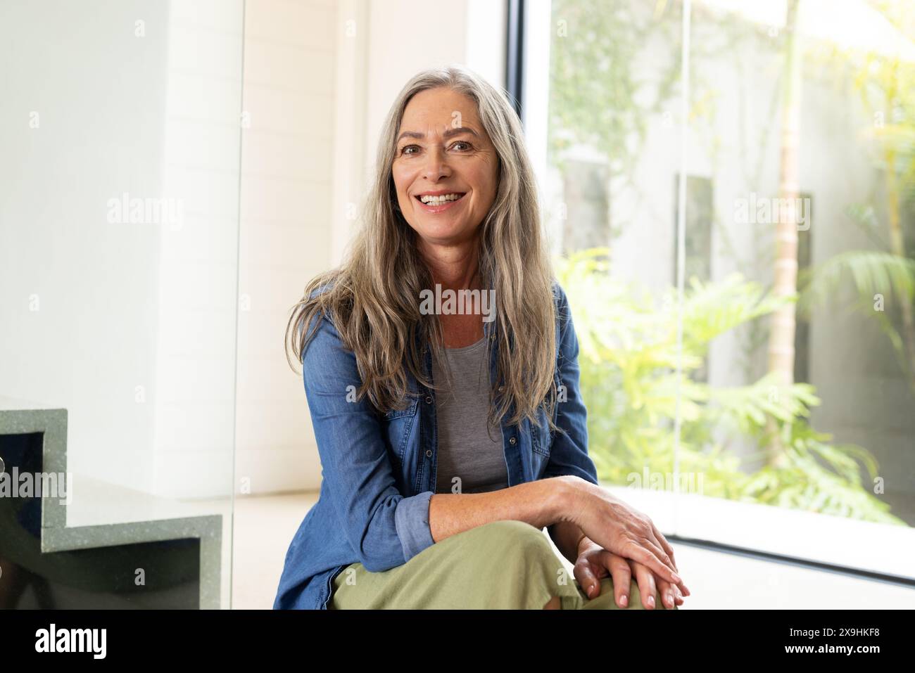A casa, donna caucasica anziana con lunghi capelli grigi e occhi blu sorridenti all'interno. Indossando una camicia blu sopra la parte superiore grigia e pantaloni verdi, seduti vicino a vincere Foto Stock