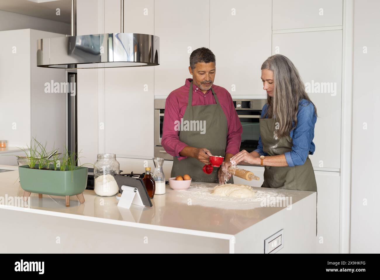 A casa, diverse coppie senior cucinano in cucina moderna, entrambe con grembiuli. Donna caucasica matura con i capelli lunghi grigi che arrotolano la pasta, biracia matura Foto Stock
