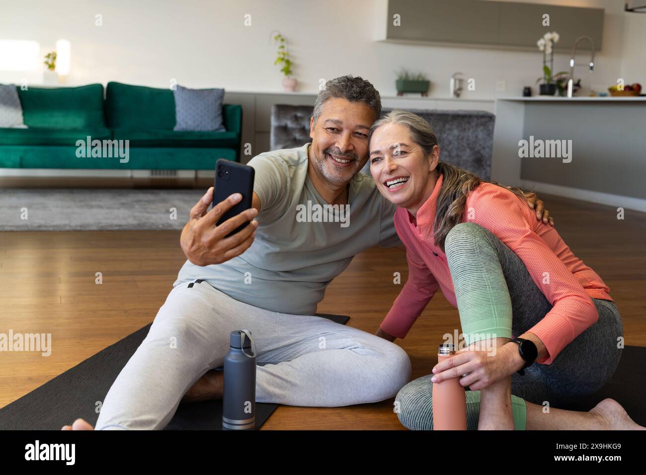 A casa, diverse coppie di anziani sorridono per un selfie sui tappetini da yoga in soggiorno. Marito birazziale con capelli ricci corti e barba grigia, moglie caucasica wi Foto Stock