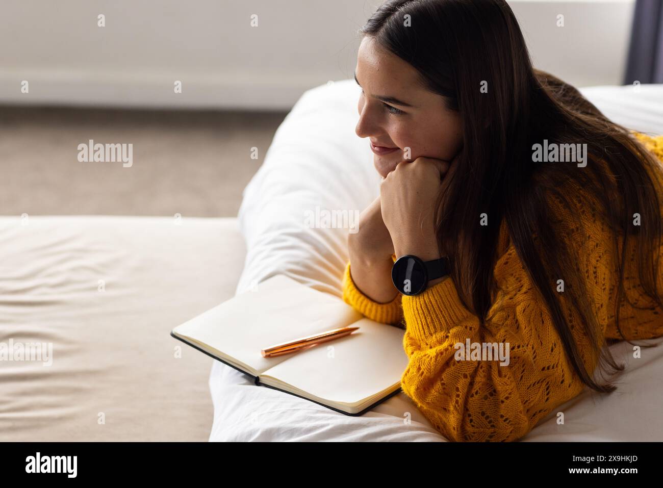 A casa, giovane donna caucasica che si rilassa sul letto con notebook e penne aperti Foto Stock