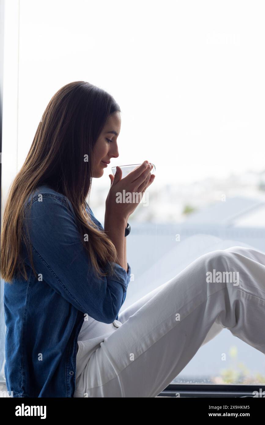 A casa, giovane donna caucasica che si gode una tazza di tè, rilassandosi vicino alla finestra Foto Stock