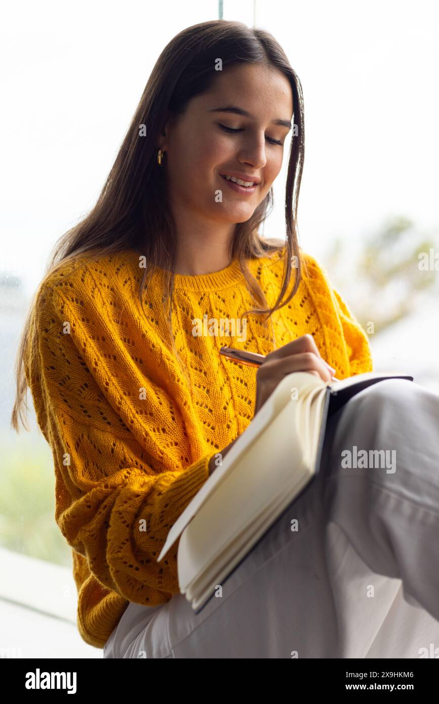 A casa, giovane donna caucasica con maglione giallo che scrive su un taccuino Foto Stock