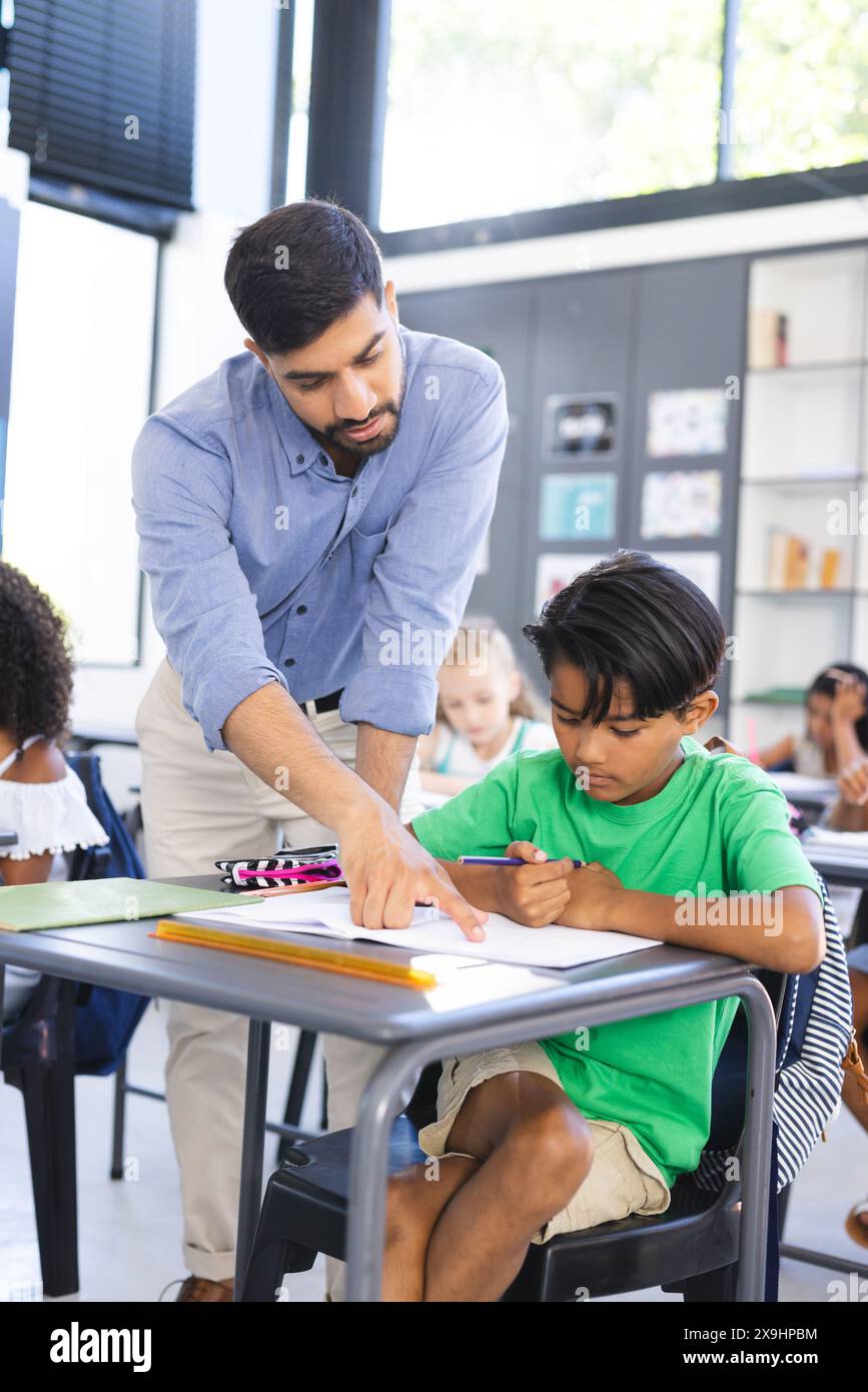 Giovane insegnante asiatico maschile assiste un ragazzo birazziale in una classe scolastica. L'insegnante si appoggia sulla scrivania, guidando la scrittura del bambino durante una lezione. Foto Stock