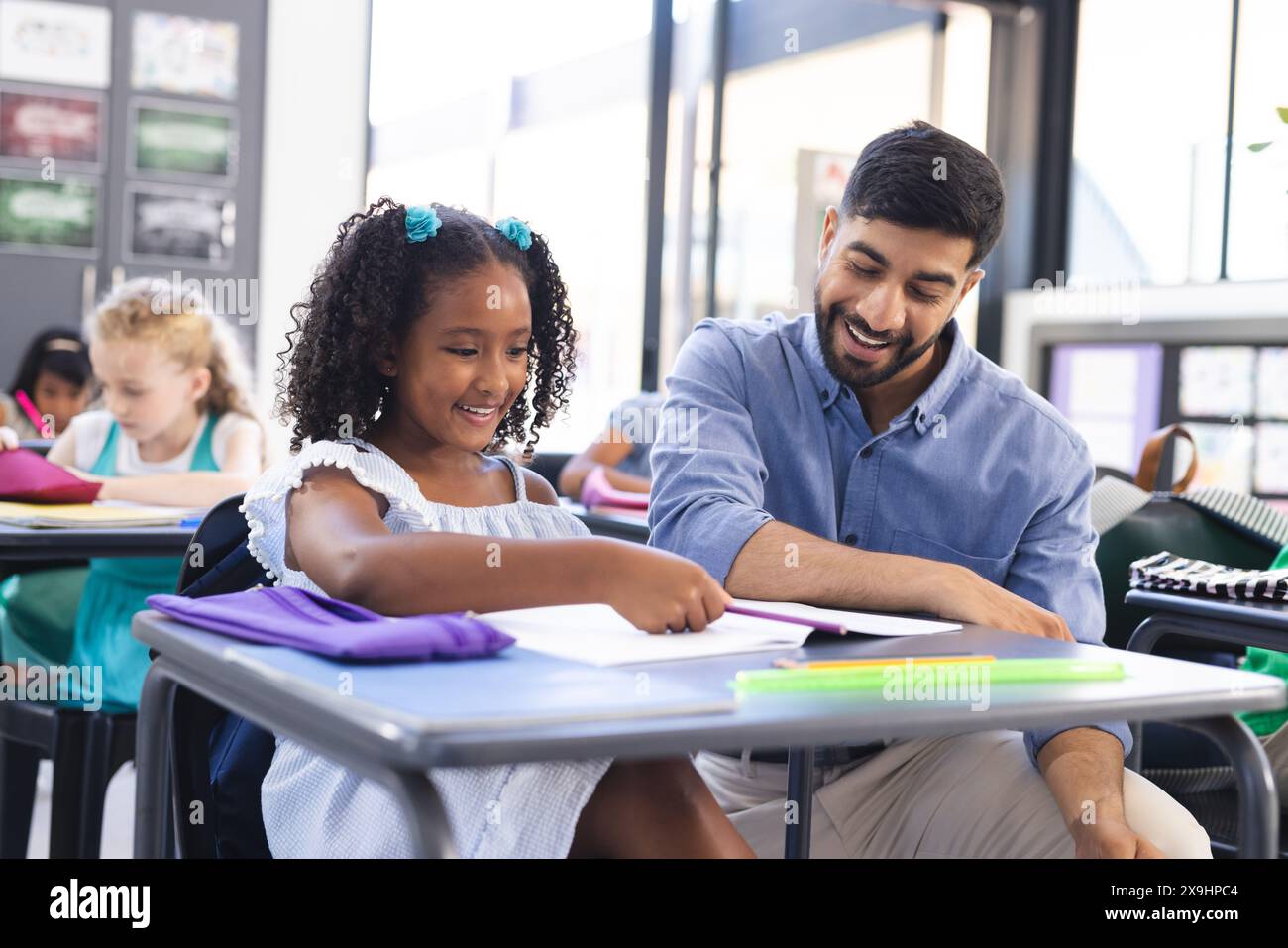 A scuola, insegnante maschile asiatico che aiuta la giovane studentessa birazziale in classe. Entrambi sono seduti a una scrivania con le forniture scolastiche, sorridendo mentre si tengono Foto Stock