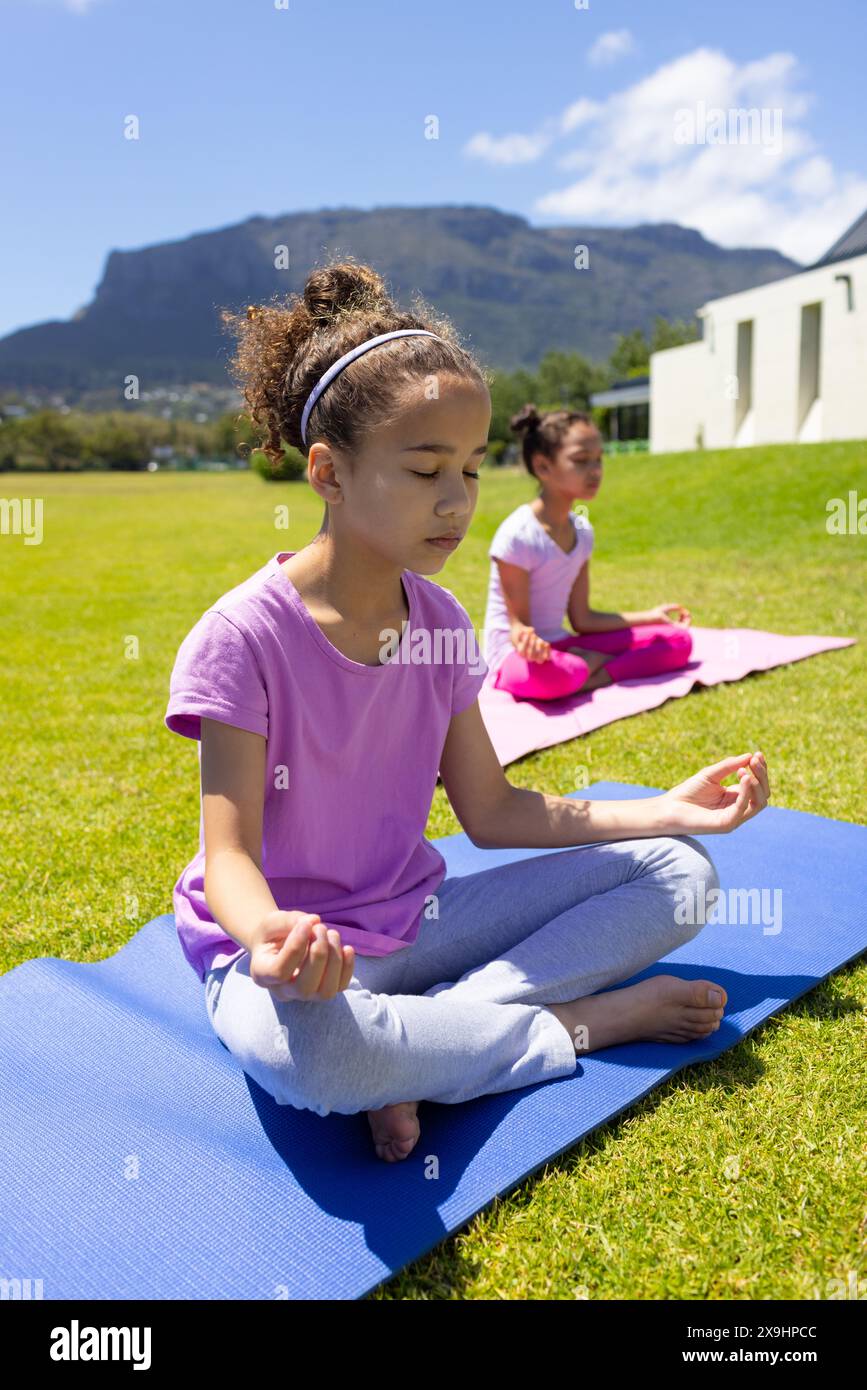 Due ragazze birazziali praticano yoga all'aperto, una con gli occhi chiusi in meditazione Foto Stock