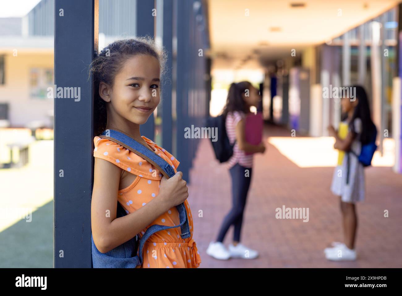 Ragazza birazziale con uno zaino sorride, in piedi in un corridoio scolastico con spazio per le copie Foto Stock