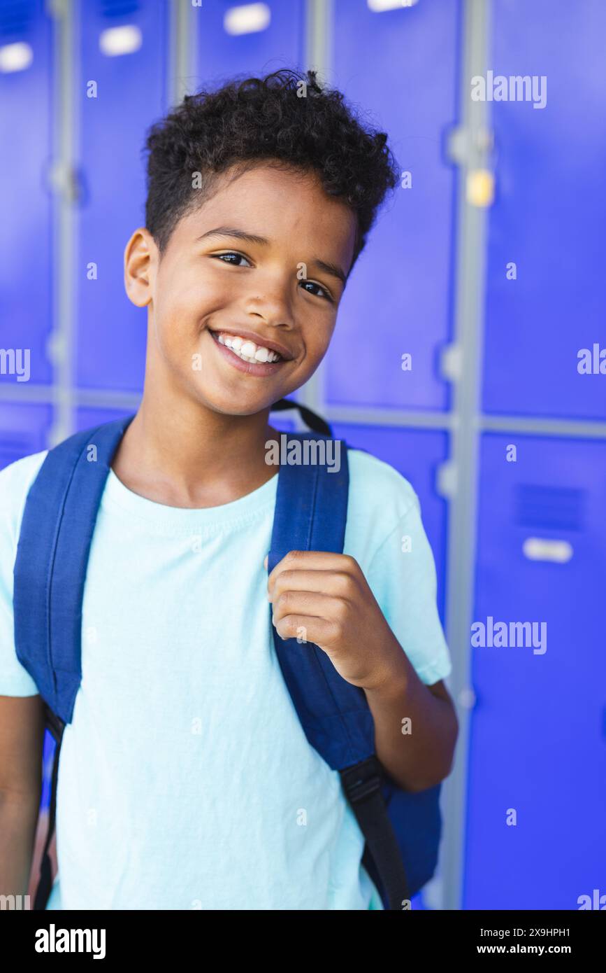 Un ragazzo birazziale con un sorriso brillante si erge davanti agli armadietti blu a scuola, indossando uno zaino Foto Stock
